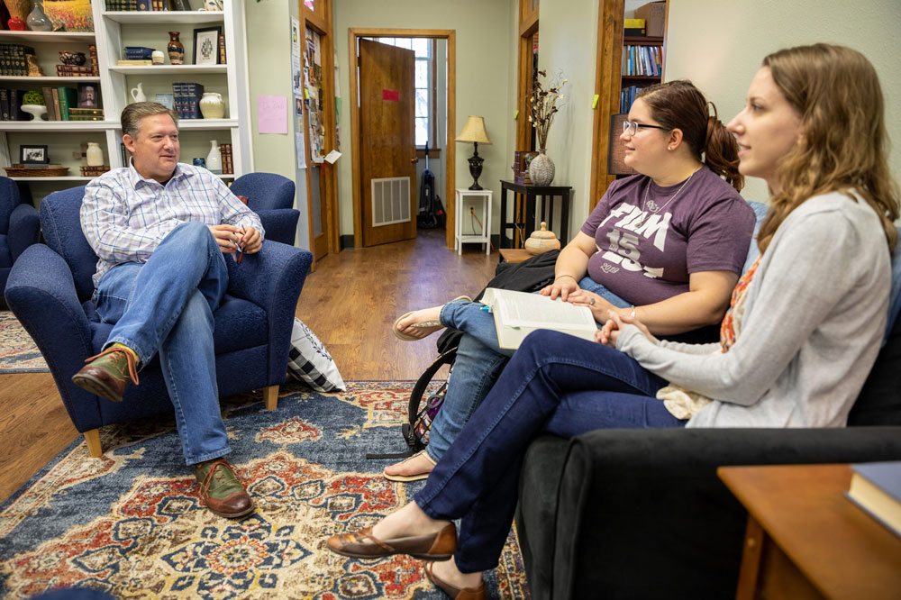 A professor sitting in an armed chair chatting with two students sitting on a couch
