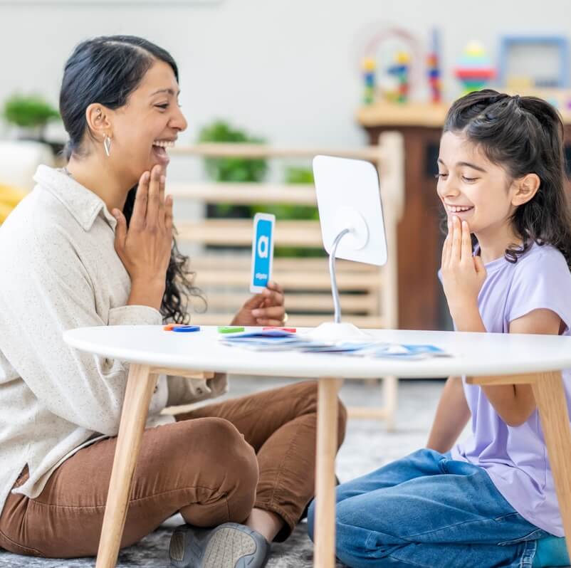 A female speech therapist works with a young girl as they practice their sounds and letter enunciation. The Therapist is dressed casually and is holding up alphabetical cue cards as they work together.