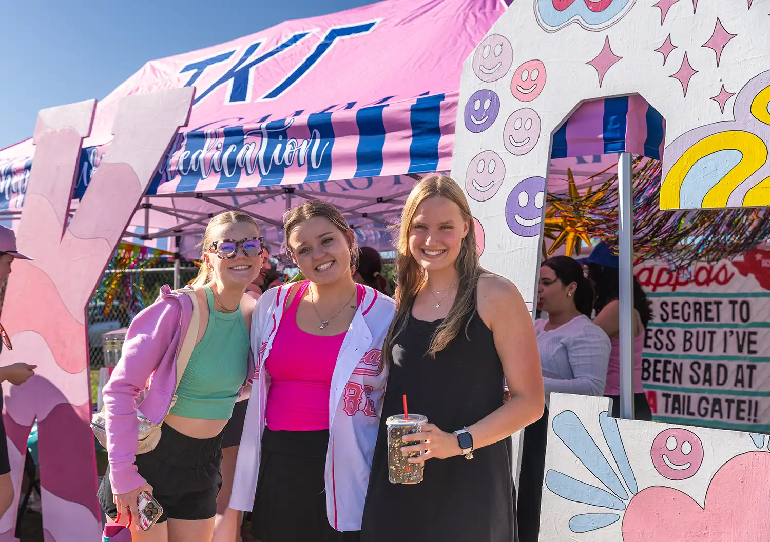 Smiling students snap a pic in front of the Tri Kappa Gamma tent at a tailgate party.
