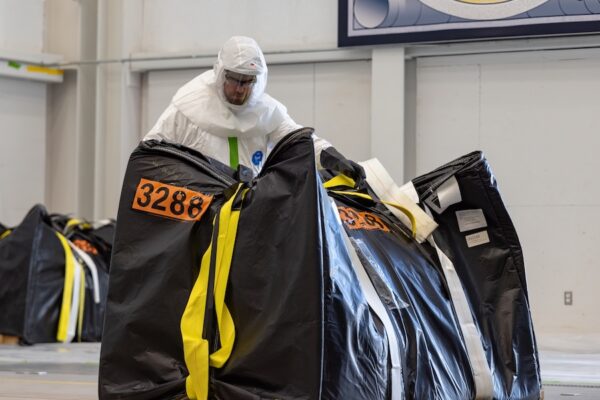 A man in personal protective gear maneuvers a large black container.