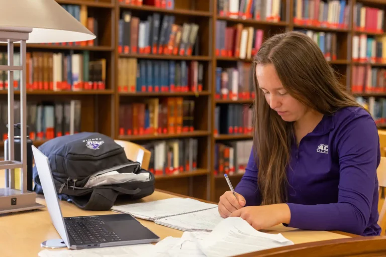 A student working alone with a laptop and papers in the library.