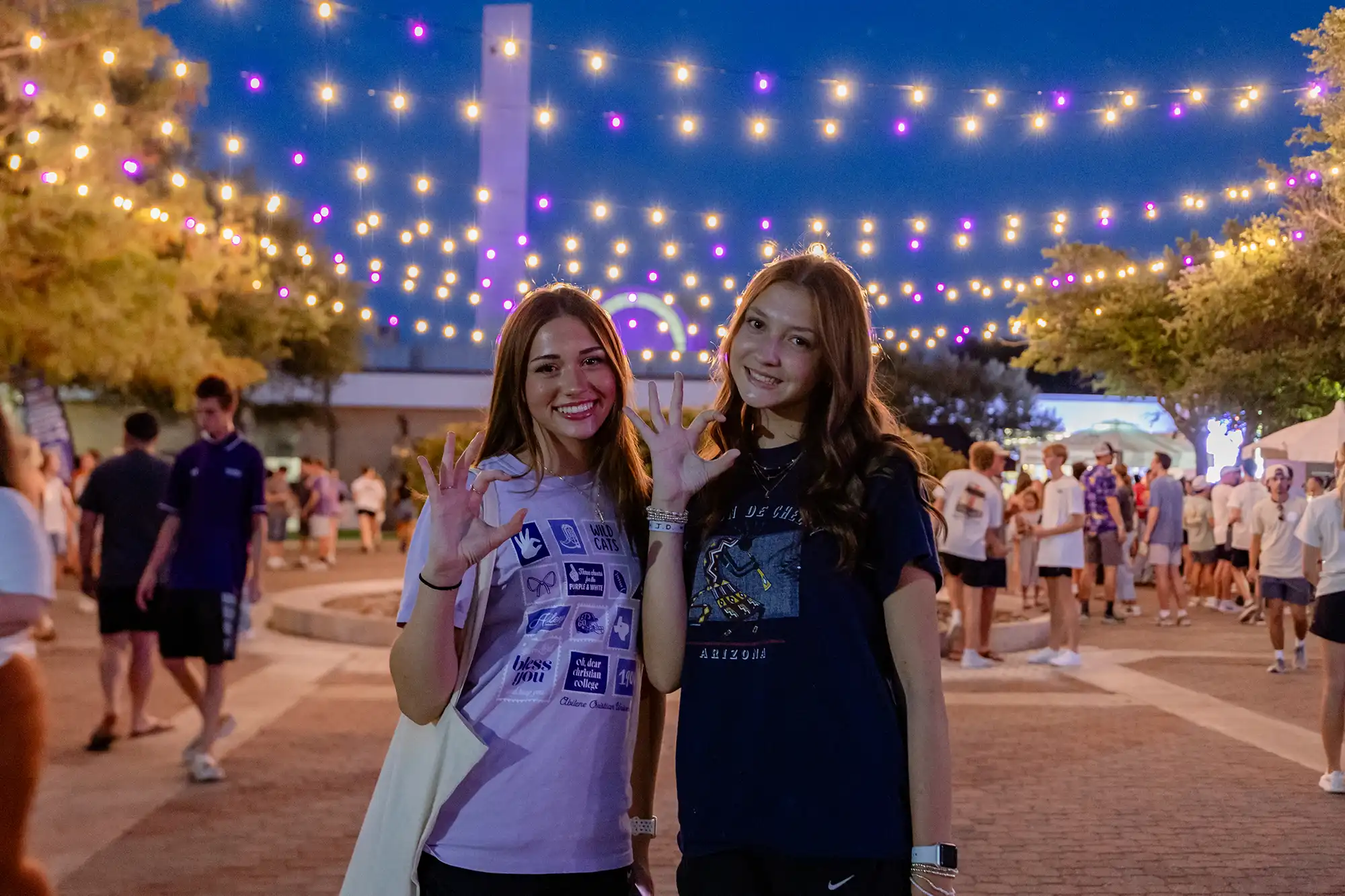 Two new Wildcats pose with hands making a W-C at an evening mixer lit by string lights in purple and white.